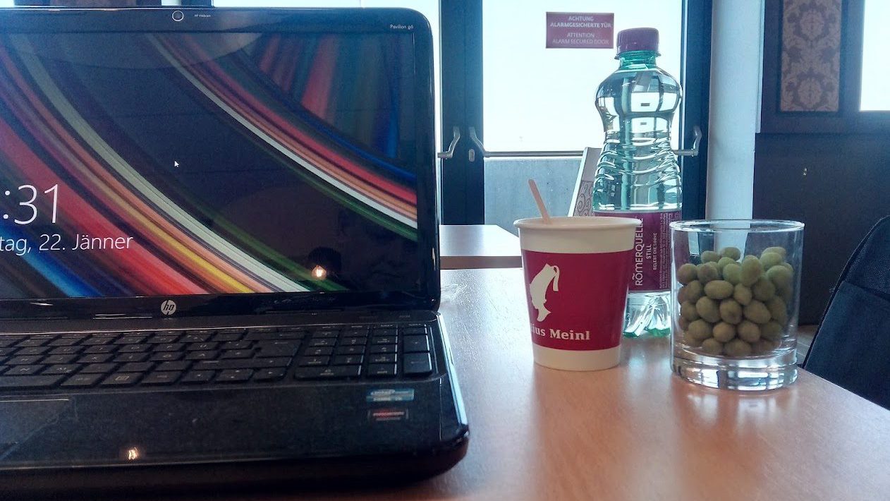 Aufgeklappter schwarzer HP-Laptop auf einem Holztisch, daneben ein Kaffeebecher, eine Wasserflasche und ein Glas mit grünen Nüssen