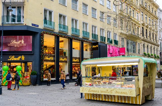 Lindt-Geschäft mit goldenen Schriftzügen und Weihnachtsgirlande an einer Fußgängerzone in Wien, davor ein Verkaufsstand mit Süßigkeiten