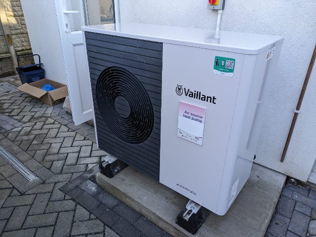 Außengerät einer Luftwärmepumpe mit großem Ventilator an einer weißen Hauswand, auf einem Betonsockel neben der Hintertür