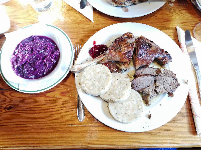 Teller mit gebratenem Gansl, drei Serviettenknödelscheiben und Preiselbeeren, daneben eine Schüssel Rotkraut auf einem Holztisch