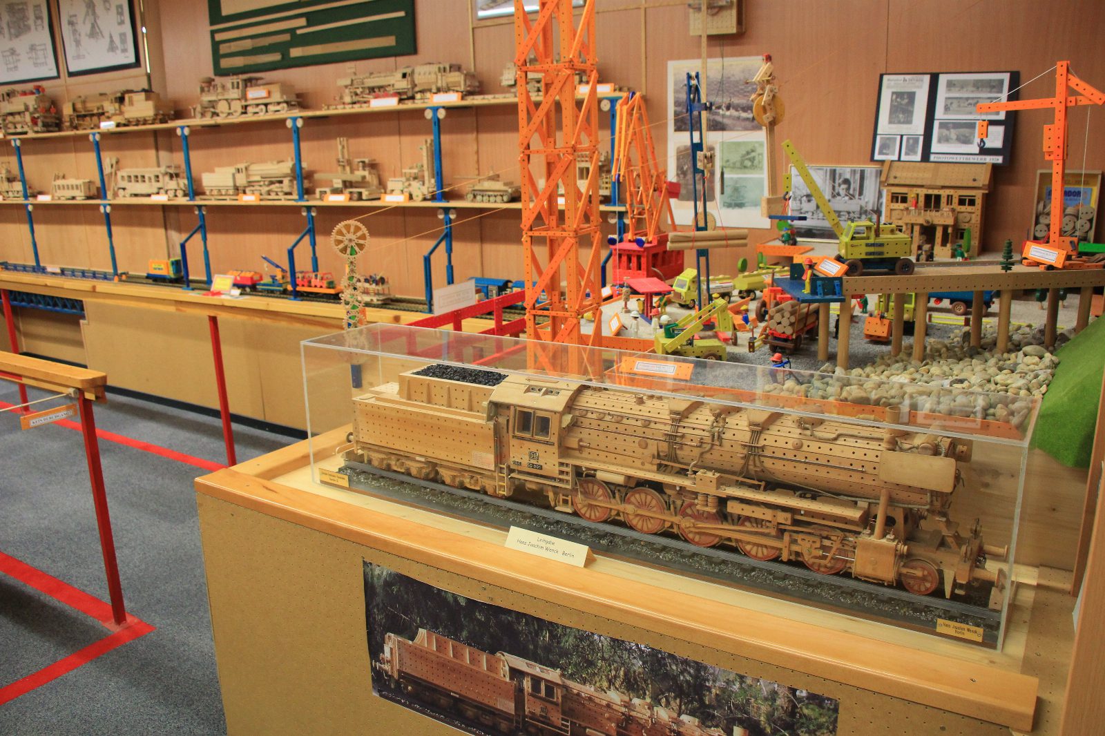 Dampflokomotive aus hellem Holzbaukasten in einer Glasvitrine, dahinter Regale mit Holzmodellen und ein orangefarbener Kran in der Matador-Ausstellung