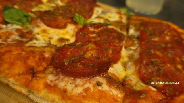 Pinsa Diavola mit scharfer Salami, geschmolzenem Fior di Latte und Basilikum auf einem Holzbrett, dahinter zwei Gläser