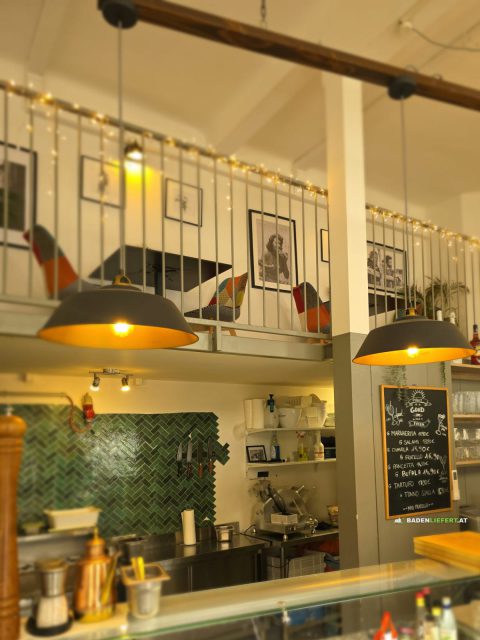 Offene Theke mit grünen Fliesen und zwei großen Hängelampen, darüber eine Galerie mit bunten Stühlen