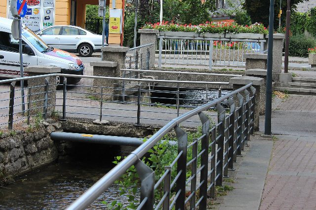 Mühlbach in Traiskirchen mit Steinmauer, Metallgeländer und einer kleinen Brücke, dahinter Blumentröge mit roten Geranien
