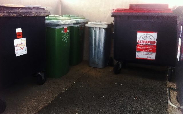 Mehrere Mülltonnen nebeneinander, darunter zwei grüne Biotonnen, eine Blechtonne und ein Altpapiercontainer mit rotem Deckel