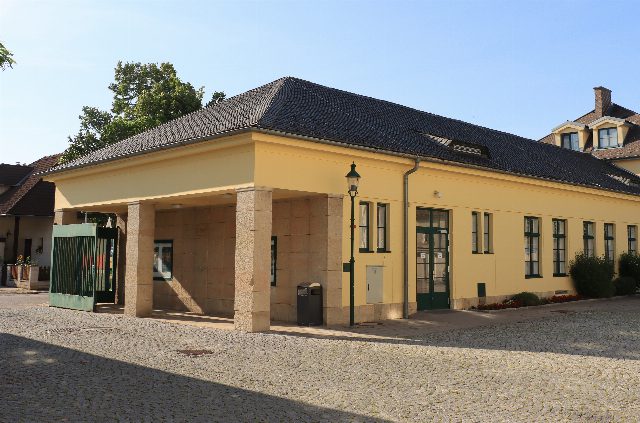 Gelbes Torgebäude mit Säulenvorbau und grünem Gittertor am Eingang zum Museum Traiskirchen, dahinter Teile der alten Fabrikgebäude