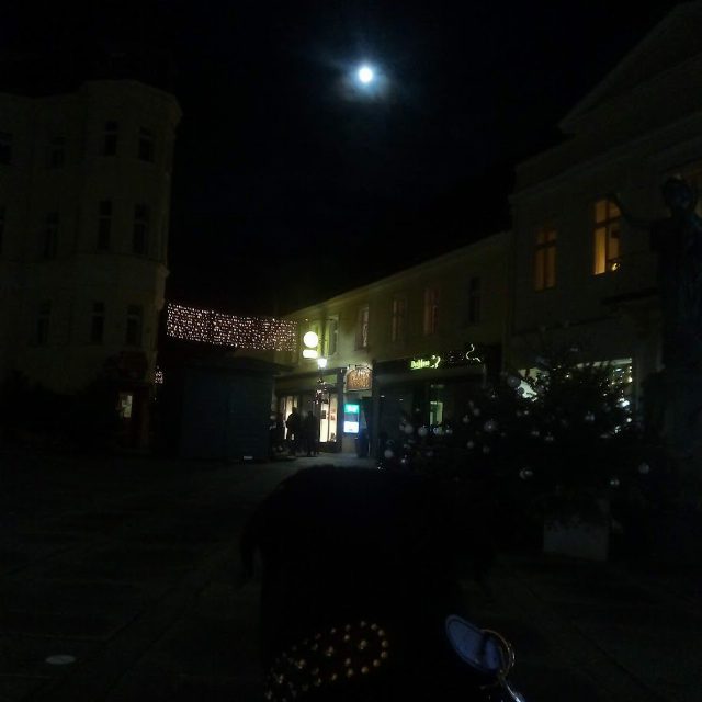 Nachtaufnahme mit Vollmond über einem Platz: beleuchtete Geschäfte, eine Lichterkette zwischen zwei Häusern und ein geschmückter Christbaum