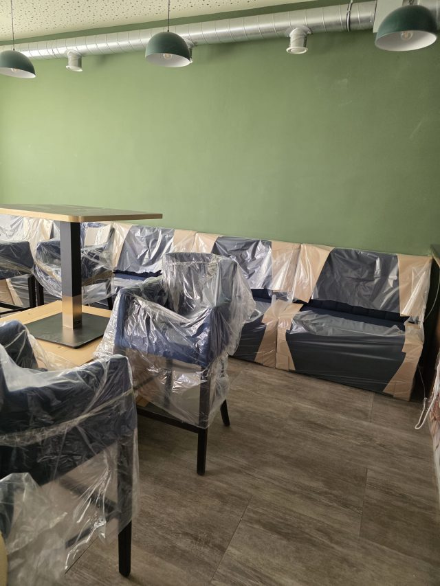Eingepackte blaue Sessel und Sitzbänke vor einer grünen Wand, darüber Lüftungsrohre und grüne Hängeleuchten