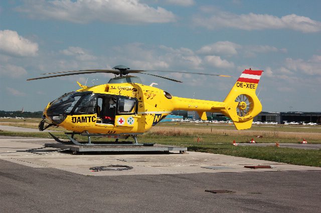 Gelber ÖAMTC-Notarzthubschrauber Christophorus 3 steht auf einem Landeplatz am Flugfeld, im Hintergrund Hangars und Kleinflugzeuge