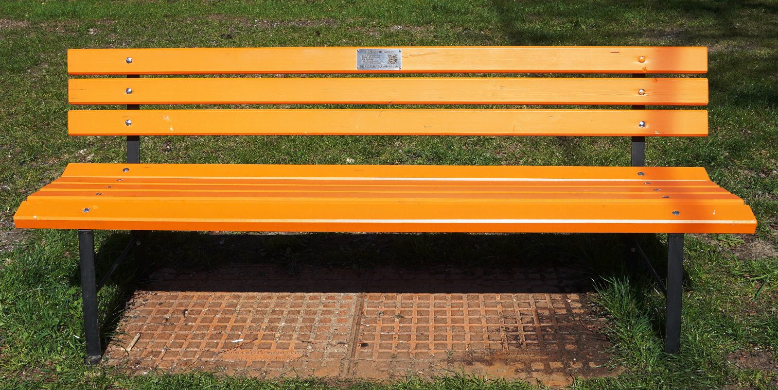 Leuchtend orange Parkbank aus Holzlatten mit kleiner Metalltafel an der Lehne, auf einer Wiese im Sonnenlicht