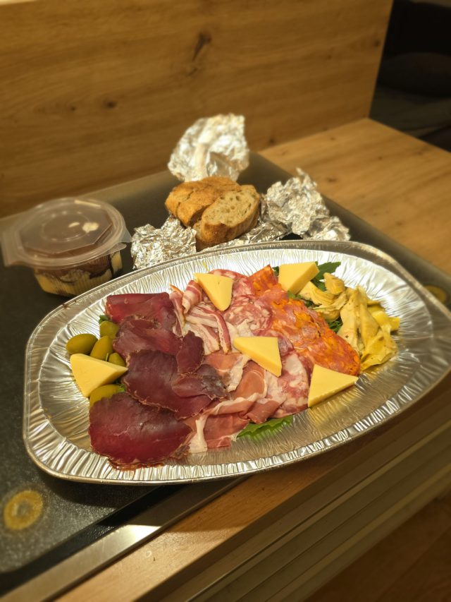 Antipasto Anima per Due auf einer ovalen Lieferplatte mit Aufschnitt, Käse, Oliven und Artischocken; dahinter Brot und Tiramisu.