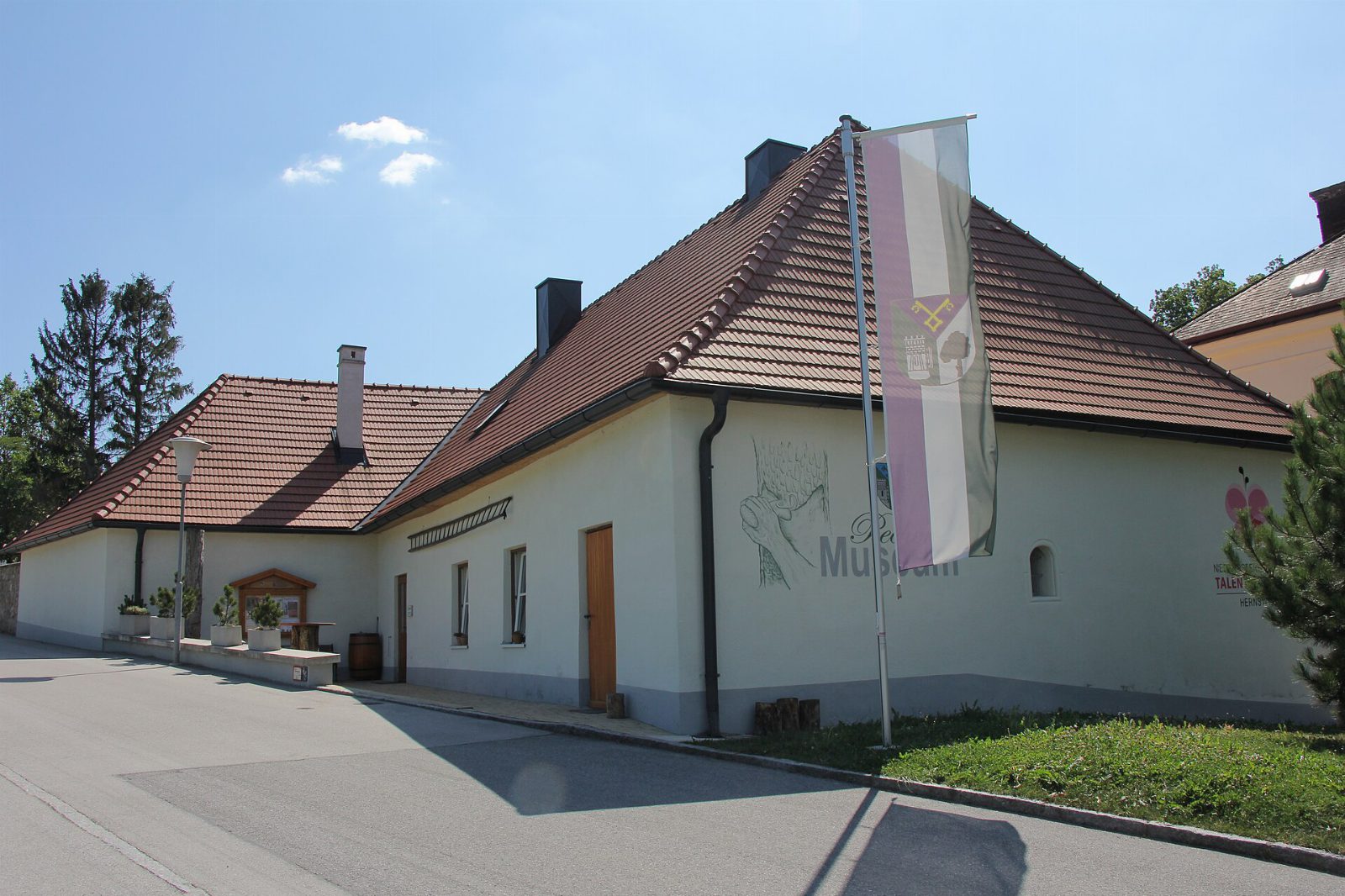 Weißes Gebäude des Pechermuseums Hernstein mit Ziegeldach, Wandbild und Gemeindefahne