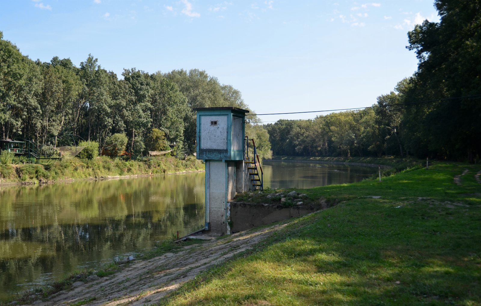 Kleines türkisgrünes Pegelhaus auf einem Betonsockel am Ufer eines ruhigen Flusses, mit Messskala am Sockel, dahinter Auwald