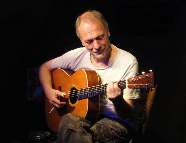 Peter Ratzenbeck sitzt mit einer akustischen Gitarre auf einer dunklen Bühne und spielt