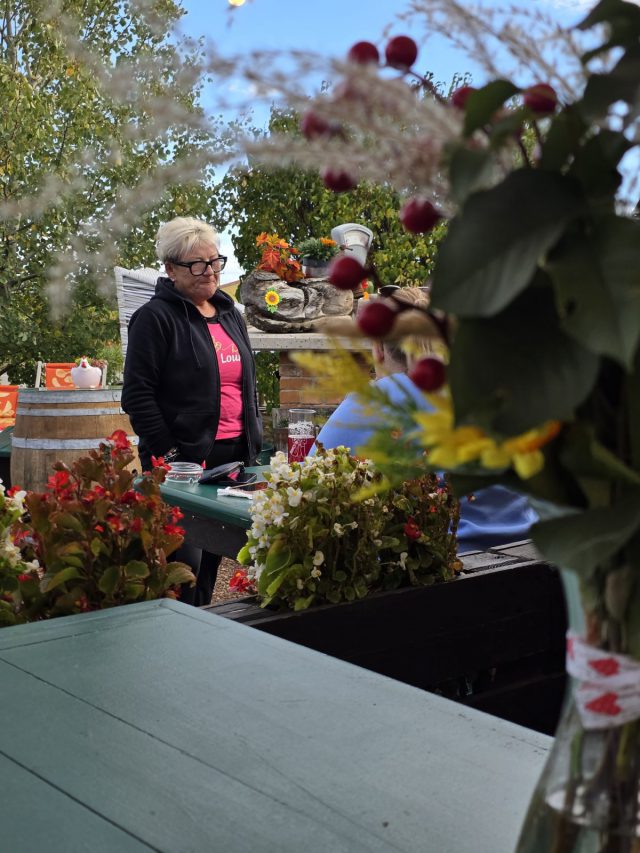 Die Wirtin der Reb Lounge im Gastgarten zwischen Blumenkästen, Weinfass und Herbstdekoration, im Vordergrund ein Blumenstrauß.