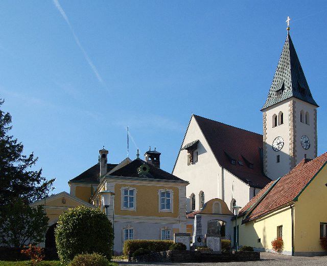 Pfarrkirche Grillenberg mit spitzem grünem Turmhelm und Uhr, links der gelbe Pfarrhof, davor ein Kriegerdenkmal, blauer Himmel