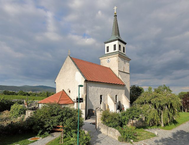 Pfarrkirche Sooß mit rotem Ziegeldach und spitzem Turmhelm, umgeben vom Friedhof, links dahinter Weingärten und der Wienerwald