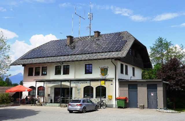 Ein zweistöckiges Gemeindeamt mit dunklen Solarmodulen auf dem ganzen Dach, davor ein Parkplatz mit Auto und ein roter Sonnenschirm