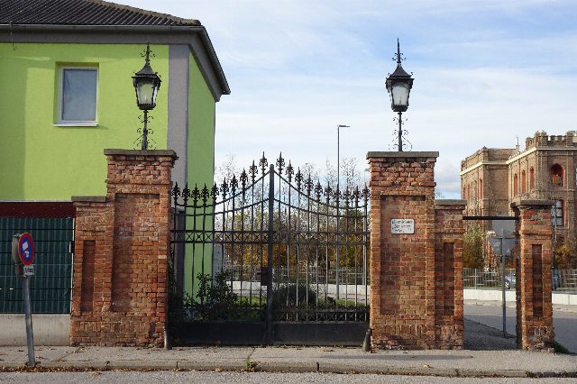 Schmiedeeisernes Tor zwischen zwei Ziegelpfeilern mit Laternen an der Alten Spinnerei Pottendorf, rechts dahinter das historische Fabrikgebäude