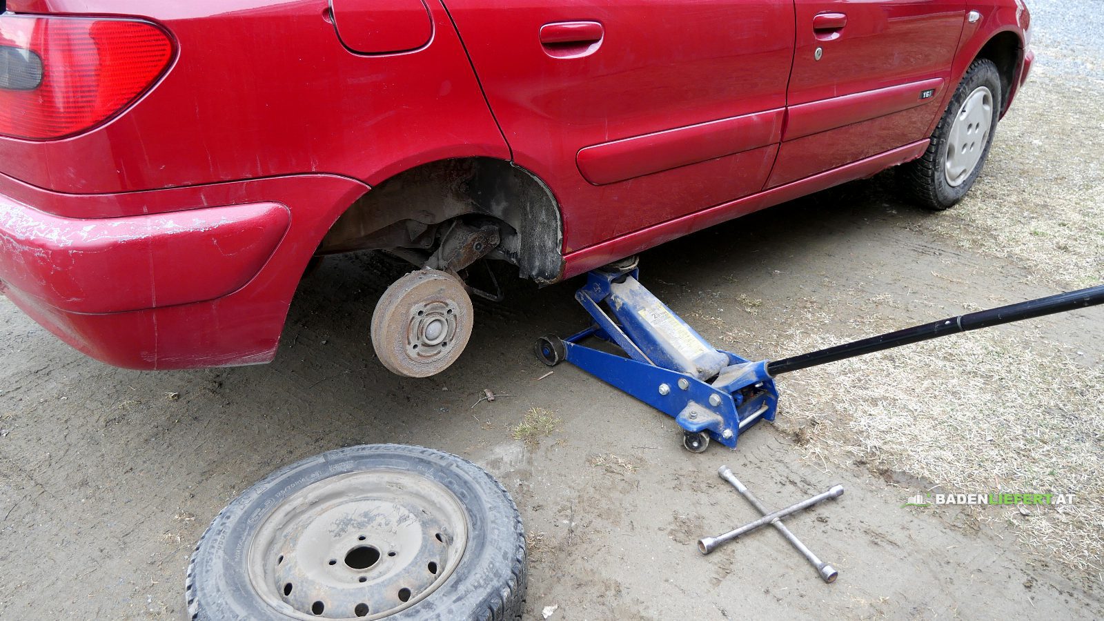 Rotes Auto auf einem blauen Rangierwagenheber, das Hinterrad ist abgenommen, davor liegen das Rad und ein Radkreuz auf dem Boden