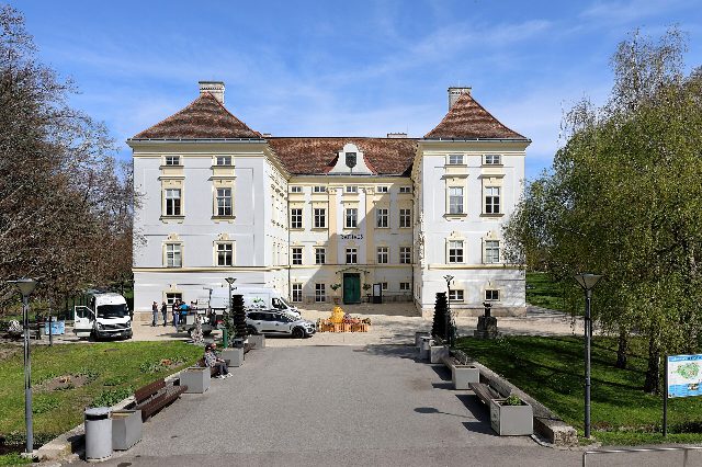 Rathaus Bad Vöslau im ehemaligen Schloss am Schlossplatz: weißer Bau mit zwei Ecktürmen und der Aufschrift Rathaus, davor Bänke und Weg im Schlosspark