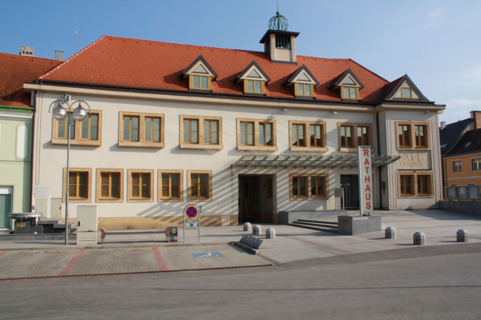 Zweigeschossiges helles Rathaus mit rotem Ziegeldach, Gauben und kleinem Uhrtürmchen, vorne ein Schild mit der Aufschrift Rathaus