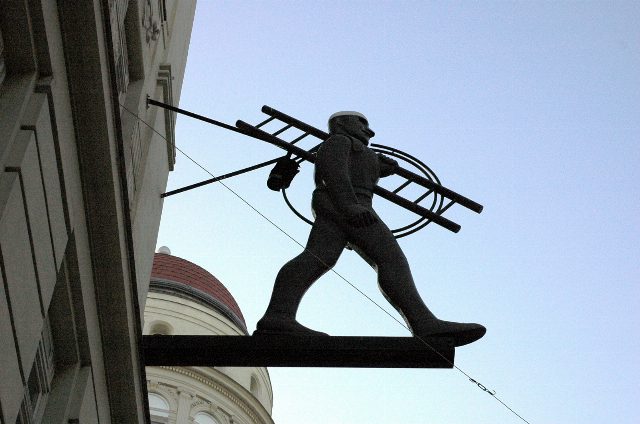 Dunkle Figur eines Rauchfangkehrers mit Leiter und Kehrseil als Ausleger an einer Hausfassade vor blauem Himmel