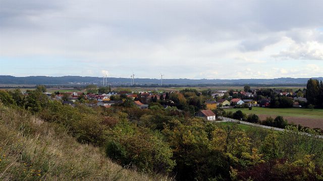 Blick über Büsche auf ein Dorf mit roten Dächern in flacher Landschaft, dahinter Windräder und Hügel unter Wolken