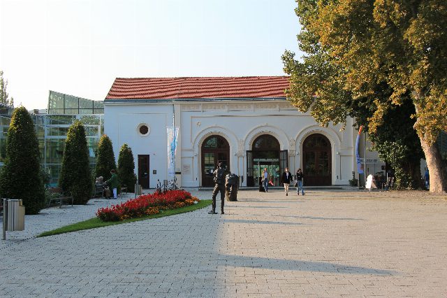 Römertherme Baden: weißer Eingangsbau mit drei Rundbogentoren und rotem Dach, links die Glashalle des Hallenbads, davor ein gepflasterter Vorplatz mit Blumenbeet