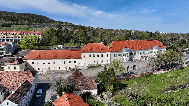 Schloss Gainfarn von oben, lang gestreckter weißer Bau mit roten Dächern, dahinter bewaldete Hügel und Weingärten