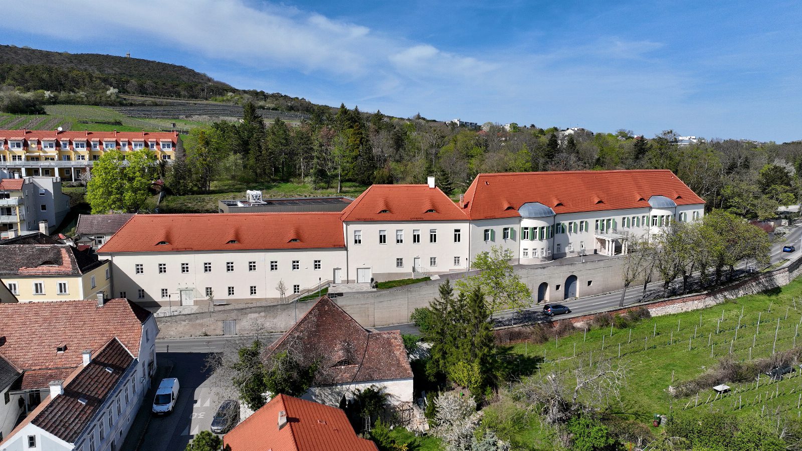 Schloss Gainfarn von oben, lang gestreckter weißer Bau mit roten Dächern, dahinter bewaldete Hügel und Weingärten
