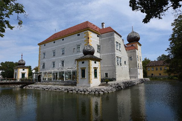 Das weiße Wasserschloss Kottingbrunn mit rotem Dach und zwei Ecktürmchen mit Zwiebelhauben, davor der Wassergraben mit Spiegelung