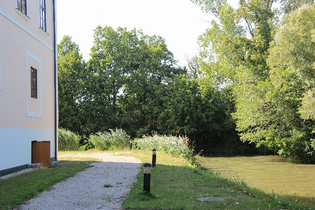 Kiesweg an der hellen Fassade von Schloss Vösendorf, daneben Wiese, Wasser und dichte Bäume im Sommer