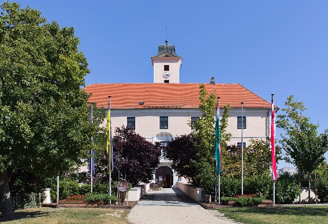 Westseite von Schloss Vösendorf mit Uhrturm, davor der Weg durch den Schlosspark zwischen Bäumen und Fahnenmasten
