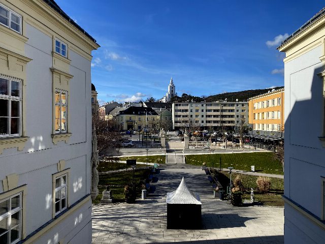 Blick vom Schloss Bad Vöslau über den Schlossplatz mit Wegen und Rasen, im Hintergrund Wohnhäuser und der Kirchturm