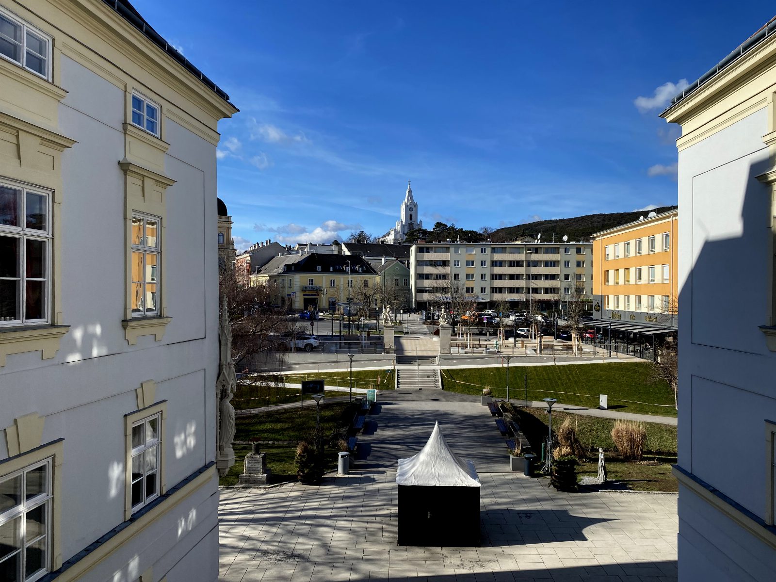 Blick vom Schloss Bad Vöslau über den Schlossplatz mit Wegen und Rasen, im Hintergrund Wohnhäuser und der Kirchturm
