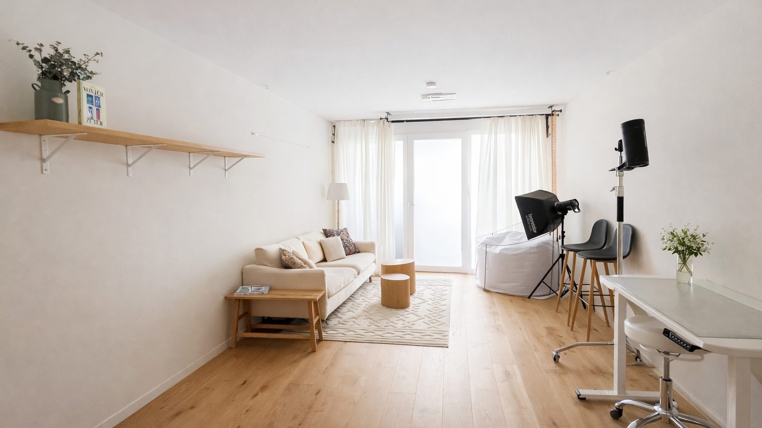 Studioraum des Singularity Studio in Baden: helles Zimmer mit Holzboden, beigem Sofa, Couchtisch aus Holz und Teppich, Wandregal mit Pflanze, rechts ein Studioblitz mit Softbox, zwei Barhocker und ein weißer Schreibtisch, hinten ein Fenster mit weißen Vorhängen