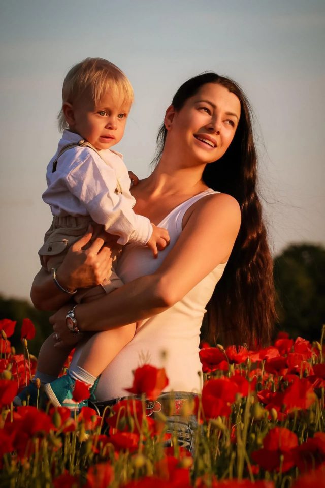 Barbora Vavro Gruber mit langen dunklen Haaren und weißem Top hält ein kleines blondes Kind auf dem Arm, beide stehen im Abendlicht in einem blühenden Mohnfeld
