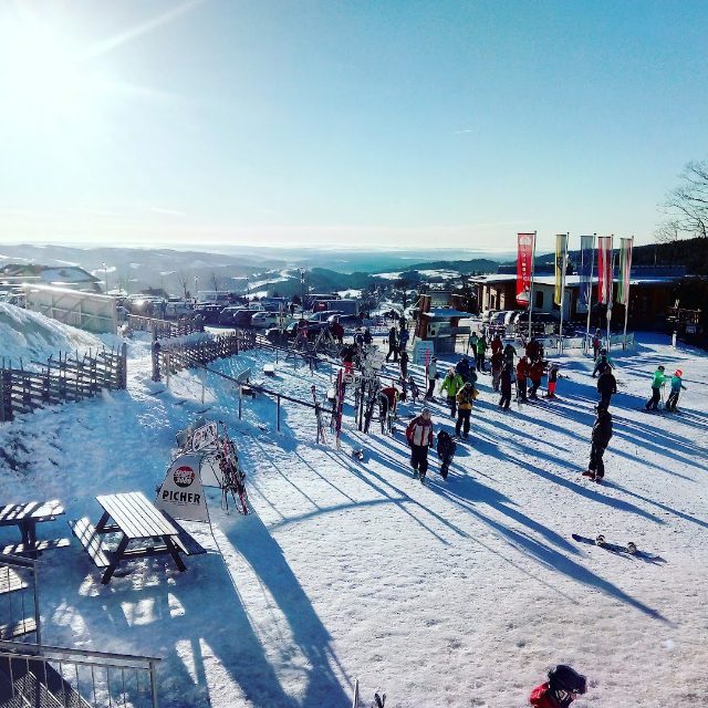 Skigebiet bei Sonnenschein: Skifahrer und Kinder im Schnee vor einem Gebäude mit Fahnen, vorne Skiständer und Holztische