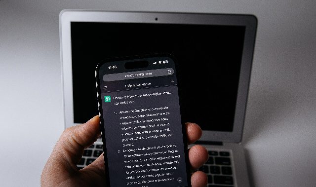 Hand hält ein Smartphone mit geöffnetem ChatGPT-Chat, dahinter ein aufgeklappter Laptop mit schwarzem Bildschirm