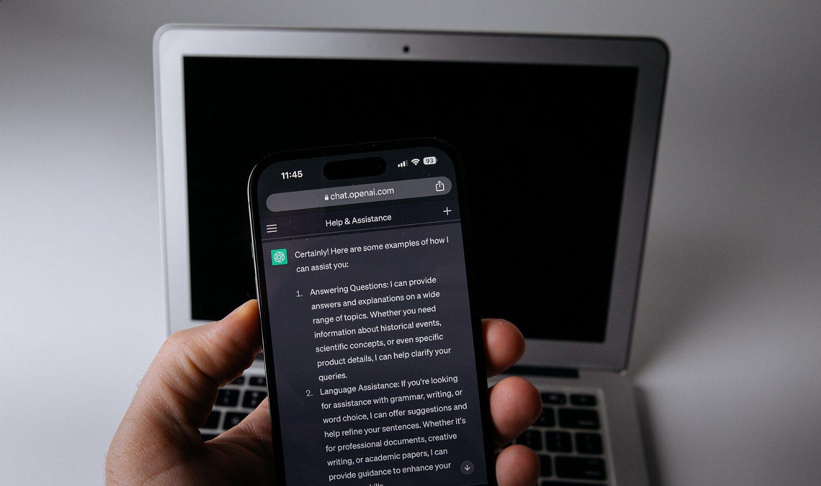Hand hält ein Smartphone mit geöffnetem ChatGPT-Chat, dahinter ein aufgeklappter Laptop mit schwarzem Bildschirm