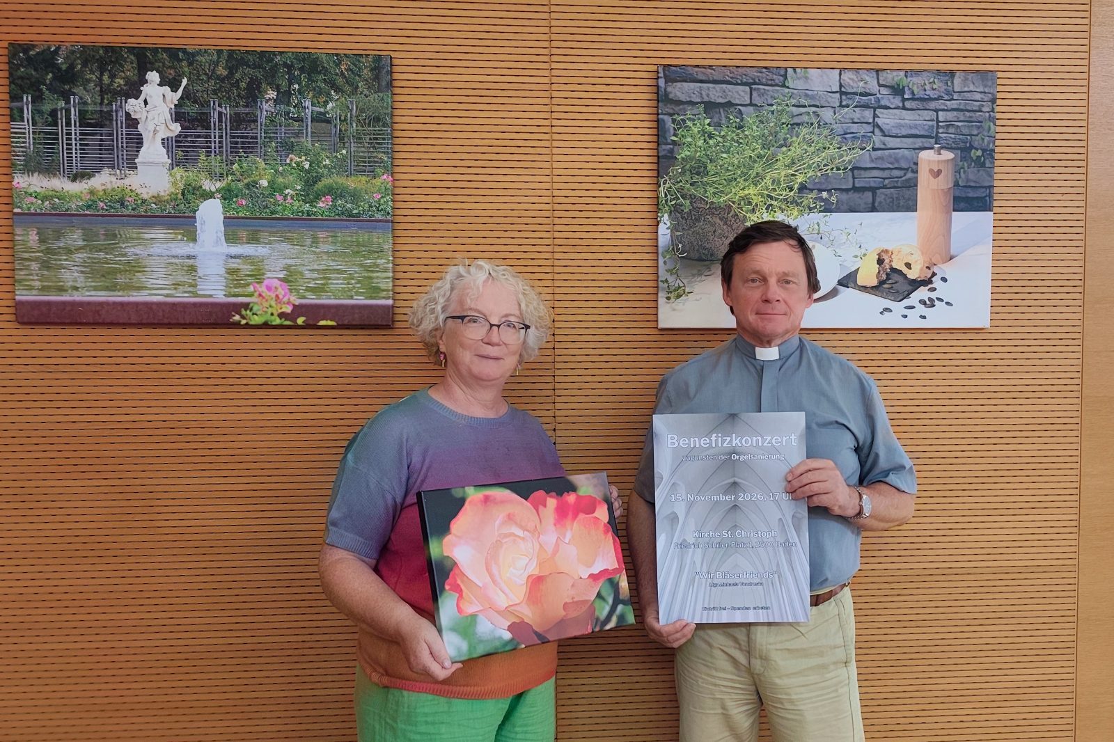 Eine Frau mit einem Leinwandfoto einer Rose und ein Geistlicher mit dem Plakat zum Benefizkonzert vor einer Holzwand mit Fotografien