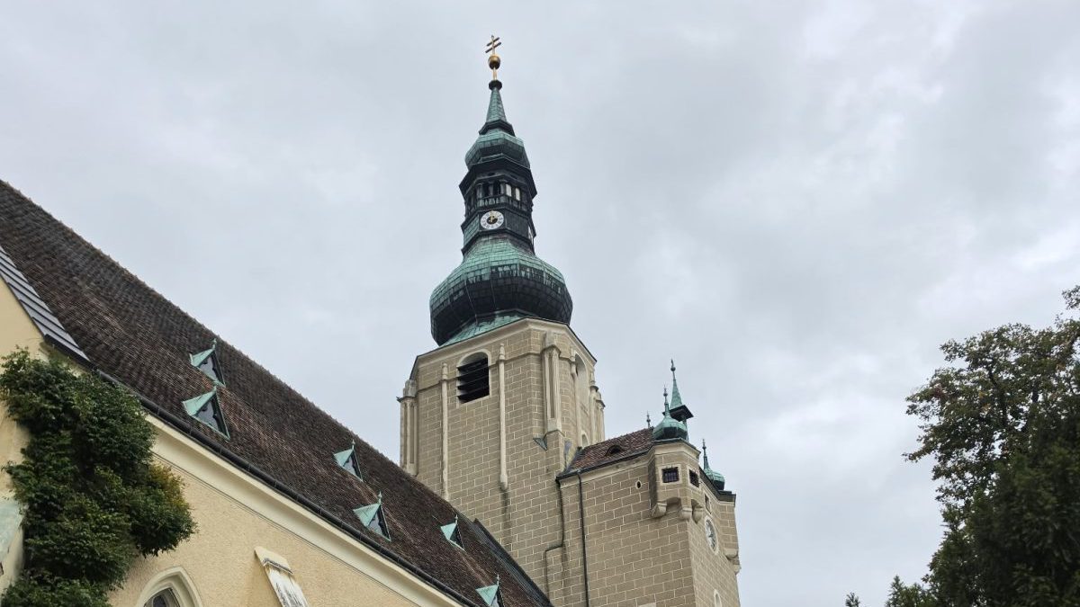 Stadtpfarrkirche St. Stephan in Baden: Turm mit grünem Zwiebelhelm, Uhr und Kreuz, links das steile Ziegeldach des Langhauses mit Efeu an der Fassade, grauer Himmel