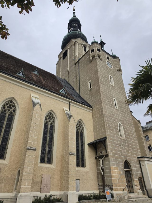 Stadtpfarrkirche St. Stephan in Baden von unten: gotisches Langhaus mit Maßwerkfenstern, daneben der Turm mit Zwiebelhelm, Uhr und einem Kruzifix an der Turmwand