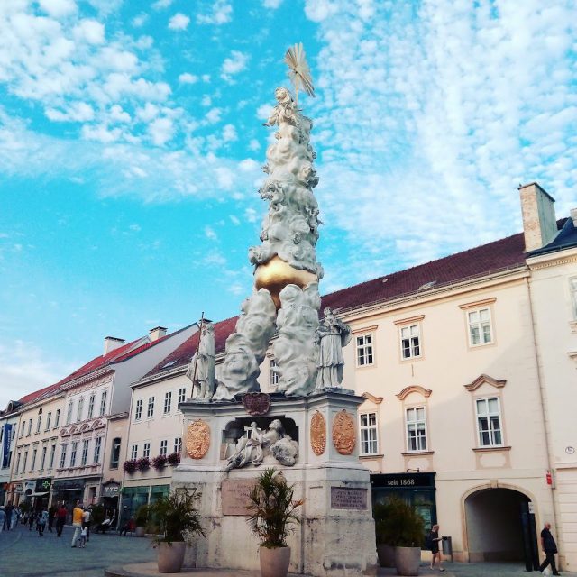 Barocke Dreifaltigkeitssäule mit vergoldeter Kugel vor hellen Bürgerhäusern, davor Passanten unter blauem Himmel mit Schäfchenwolken