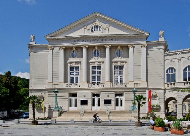 Fassade des Stadttheaters Baden mit Säulen, Giebel und der Inschrift Jubilaeums-Stadt-Theater, davor eine Freitreppe und Palmen in Töpfen