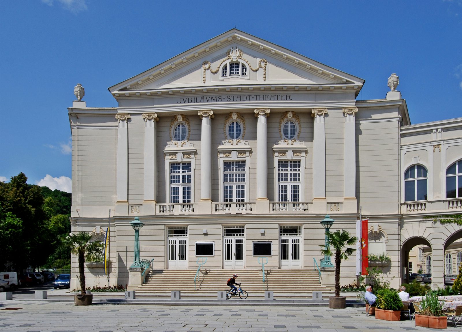 Fassade des Stadttheaters Baden mit Säulen, Giebel und der Inschrift Jubilaeums-Stadt-Theater, davor eine Freitreppe und Palmen in Töpfen