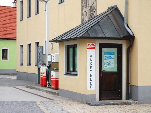 Kleine Dorftankstelle mit zwei roten Zapfsäulen und einem Kassenhäuschen mit senkrechtem Schriftzug Tankstelle an einer gelben Hauswand