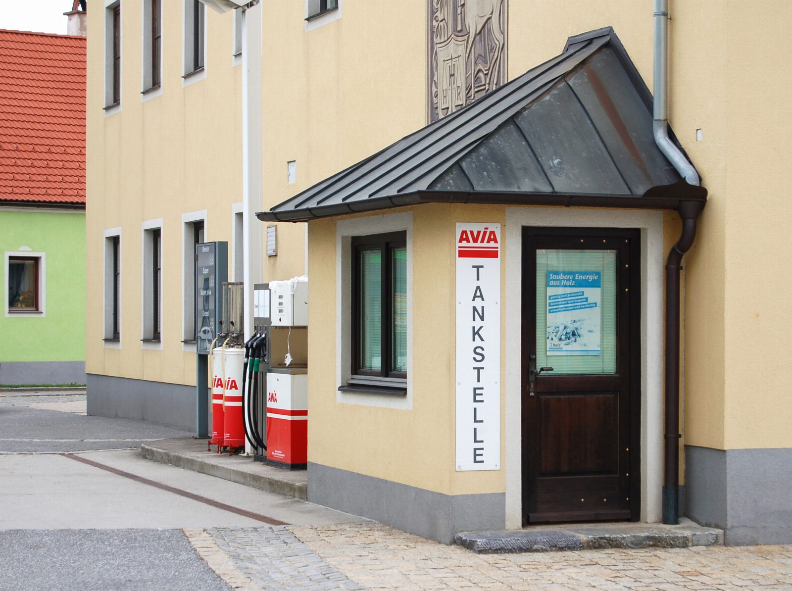 Kleine Dorftankstelle mit zwei roten Zapfsäulen und einem Kassenhäuschen mit senkrechtem Schriftzug Tankstelle an einer gelben Hauswand