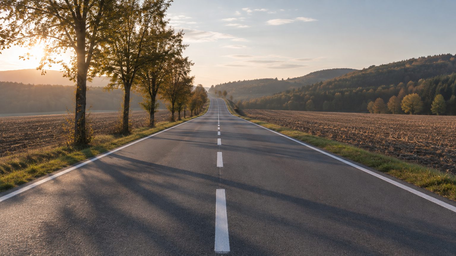 Symbolbild: leere zweispurige Landstraße im Oktobermorgenlicht, links eine Baumreihe mit langen Schatten quer über den Asphalt, rechts abgeerntete Felder, im Hintergrund bewaldete Hügel